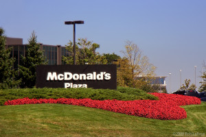 A large black sign with white letters reads McDonalds Plaza, surrounded by green grass, red flowers, and trees, with office buildings visible in the background under a clear blue sky.